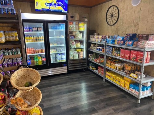 A small store interior featuring shelves of groceries, a refrigerator with drinks, and a TV displaying a basketball game.
