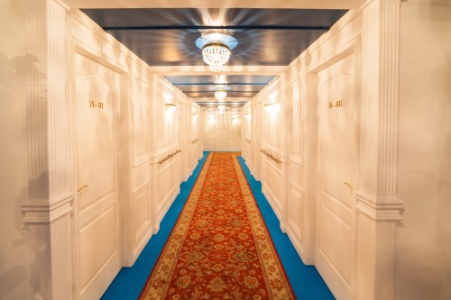 A well-lit hotel corridor with white doors, a patterned carpet, and a chandelier overhead.