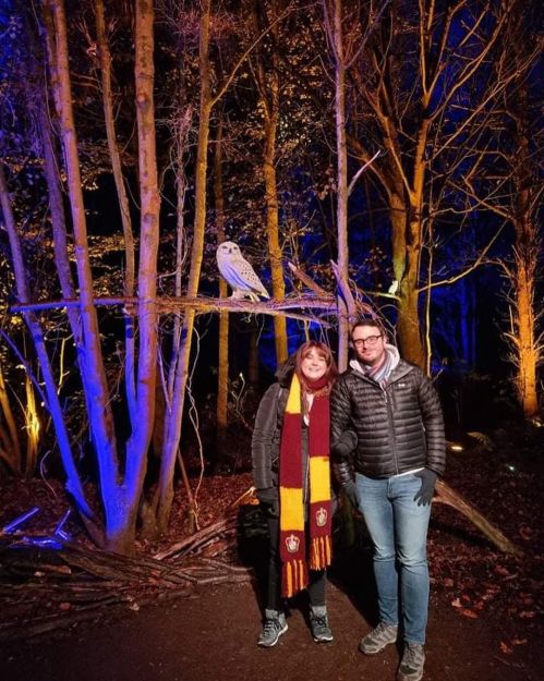 A woman and a man stand together in a forest at night, with an owl perched on a branch above them.