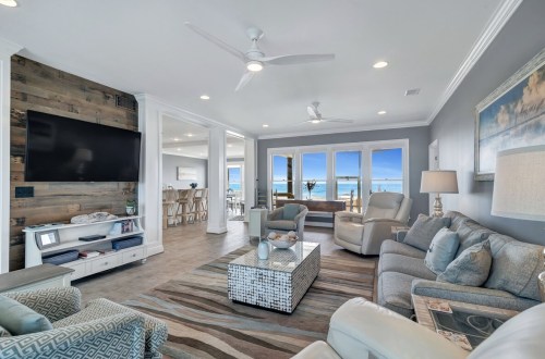 Modern living room with coastal decor, large windows, and a view of the beach. Light colors and comfortable seating.