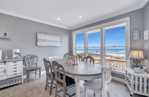 Bright dining area with a large table, chairs, and ocean views through large windows. Beach visible outside.