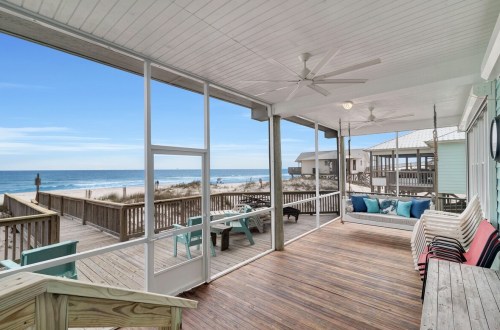 A spacious beachfront porch with seating, overlooking the ocean and sandy shore under a clear blue sky.