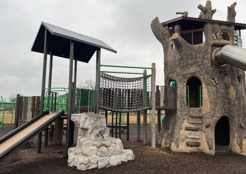 A playground featuring a treehouse structure, slide, climbing net, and a ramp, set against a cloudy sky.