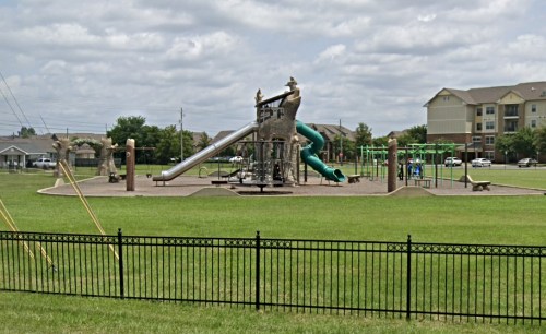 A playground with slides and climbing structures, surrounded by green grass and a black fence, under a cloudy sky.
