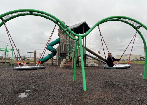A playground with green swings and a slide, featuring a child swinging on one of the swings under a cloudy sky.
