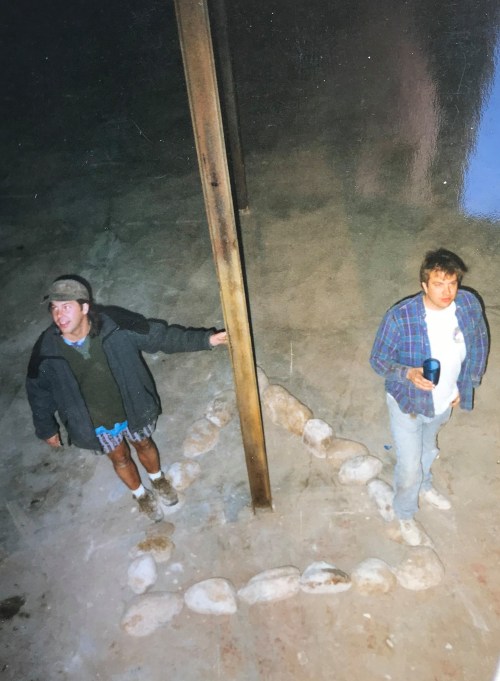 Two people stand in a dimly lit space, one holding a drink, near a stone arrangement on the ground.