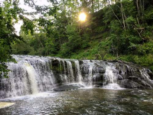 A serene waterfall cascades over rocks, surrounded by lush greenery and illuminated by the warm glow of the setting sun.