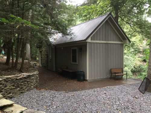 A small gray cabin nestled among trees, with a stone path and benches outside, surrounded by a natural landscape.