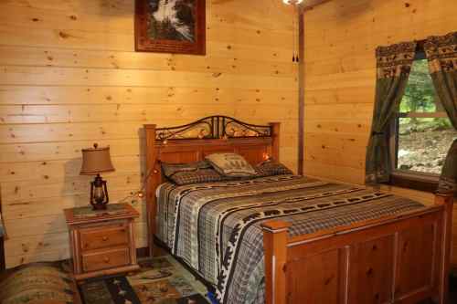 Cozy wooden bedroom with a bed, nightstand, lamp, and curtains, featuring rustic decor and a view of nature outside.
