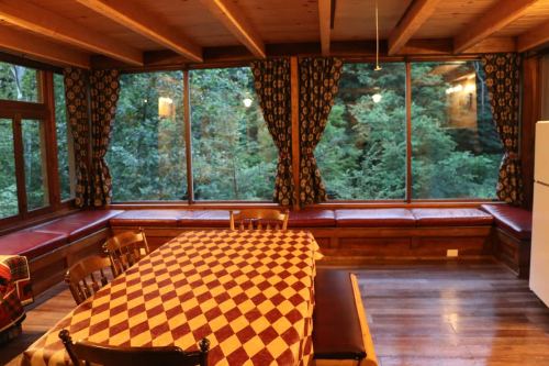Cozy wooden dining area with a checkered tablecloth, surrounded by large windows overlooking a forest.