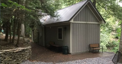 A small gray cabin surrounded by trees, with a stone wall and two benches outside on a gravel path.