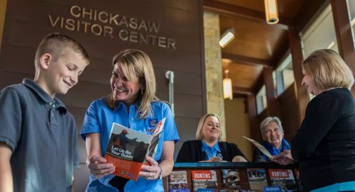 A smiling staff member assists a young boy at the Chickasaw Visitor Center, with brochures and other staff in the background.