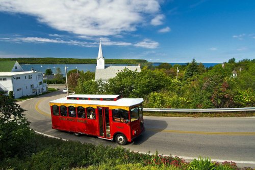 A red trolley bus drives along a scenic road with a church and lush greenery overlooking a calm body of water.