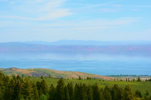 A serene landscape featuring a calm lake surrounded by rolling hills and distant mountains under a clear blue sky.