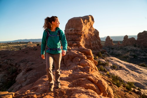 A hiker stands on a rocky outcrop at sunset, surrounded by desert landscape and unique rock formations.