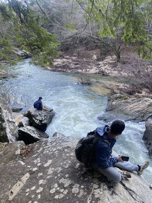 Two people sit on rocky terrain by a flowing river, surrounded by trees and a natural landscape.