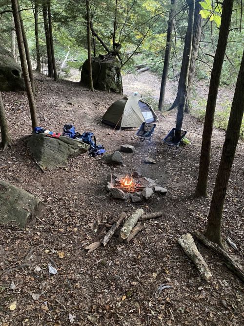 A campsite in the woods with a tent, chairs, and a fire pit surrounded by trees and rocks.