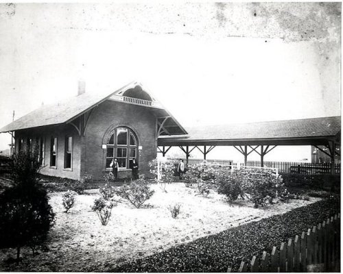 Historic train station with a brick structure, surrounded by a garden and a covered platform. Black and white photo.