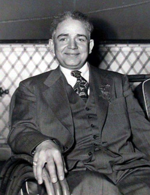 A smiling man in a suit sits comfortably in a chair, with a patterned background behind him.