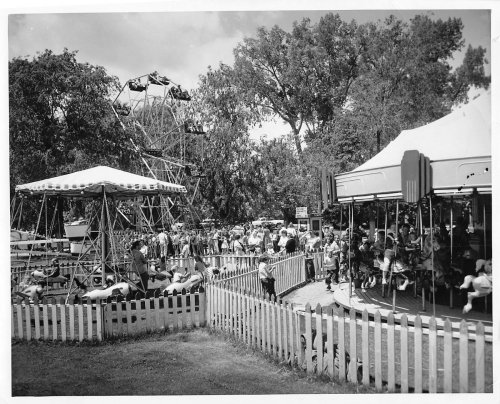 A vintage black-and-white photo of a carnival with a Ferris wheel, carousel, and crowds enjoying the attractions.