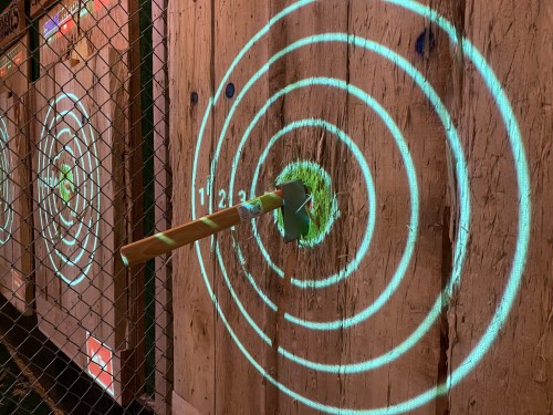 A wooden target board with concentric circles and an axe embedded in the center, illuminated by green light.