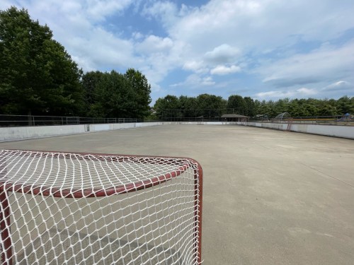 An empty outdoor hockey rink with a goal in the foreground and trees in the background under a partly cloudy sky.