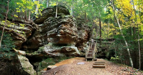 A wooden staircase leads through a forested area with colorful autumn leaves and rocky formations.