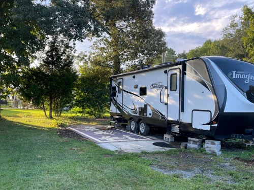 A white RV parked on a concrete pad surrounded by trees and grass, with a blue sky in the background.