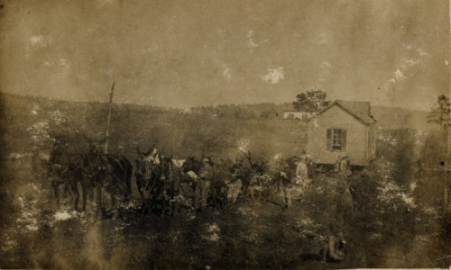 A faded historical photo of a group of people with horses near a small house in a rural landscape.