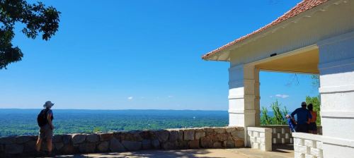 A scenic overlook with a stone wall, featuring people enjoying a panoramic view of green hills under a clear blue sky.