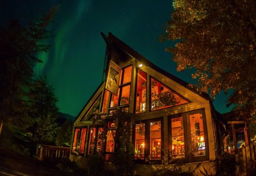 A cozy lodge illuminated at night, surrounded by trees, with the Northern Lights glowing in the sky above.