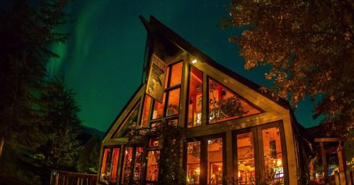 A cozy cabin illuminated at night, surrounded by trees and under a vibrant display of the northern lights.
