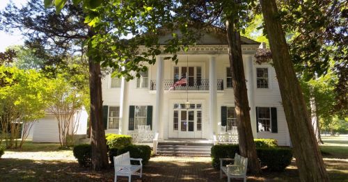 A historic white house with columns, green shutters, and an American flag, surrounded by trees and benches.