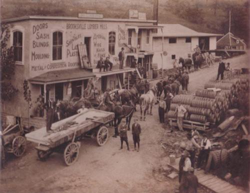 Historic black-and-white photo of a lumber mill with horse-drawn wagons, workers, and stacked lumber.