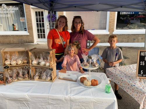 Two women and two children stand behind a table at a market, showcasing baked goods in wooden boxes and on plates.