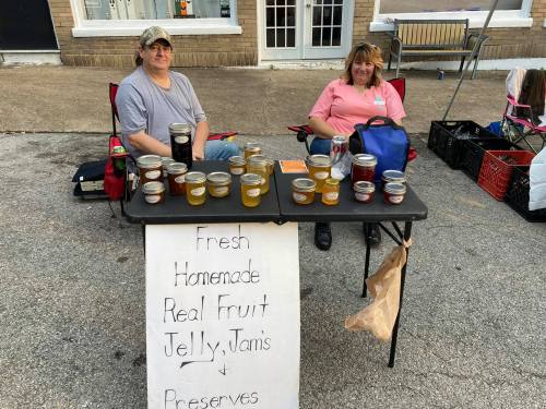 Two vendors sit at a table displaying jars of homemade fruit jelly, jams, and preserves, with a sign promoting their products.