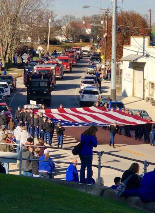 A parade scene with people holding a large American flag, surrounded by vehicles and onlookers.