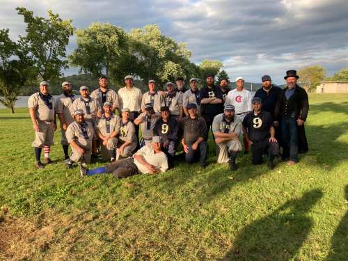 A group of 20+ people in vintage baseball uniforms poses together on a grassy field with trees in the background.
