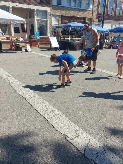 A child bends down to pick something up on a street during a market event, with adults and tents in the background.