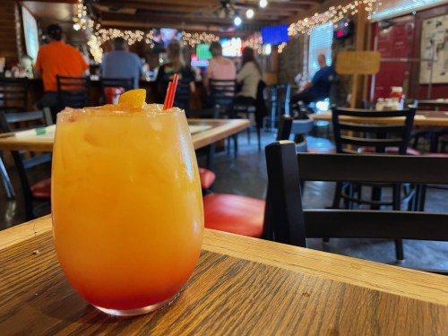 A colorful cocktail sits on a wooden table, with a lively restaurant scene in the background.