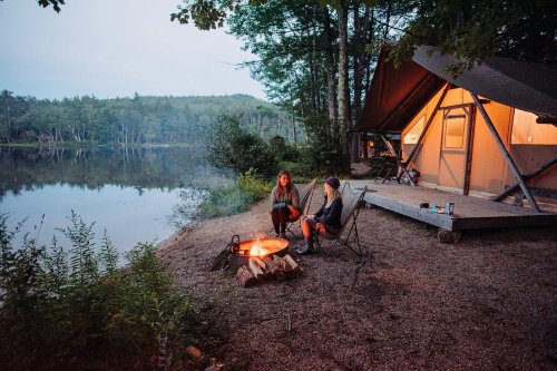 Two people sit by a campfire near a tent by a calm lake, surrounded by trees at dusk.