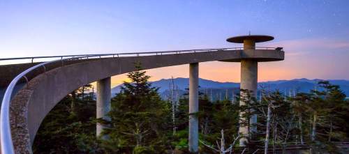 A scenic overlook bridge with a circular platform, surrounded by trees and mountains at sunset.