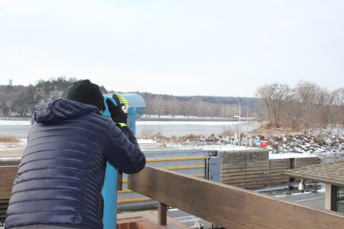 A person in a winter jacket looks through a telescope at a snowy landscape by a river.