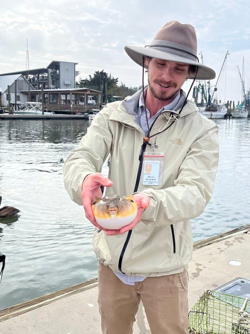 A person in a hat holds a fish near a marina, with boats and a cloudy sky in the background.