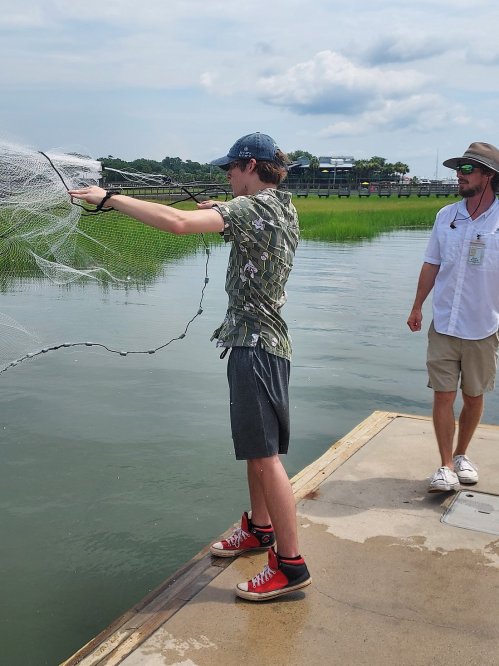 A young man casts a fishing net into the water while another man observes nearby on a sunny day.