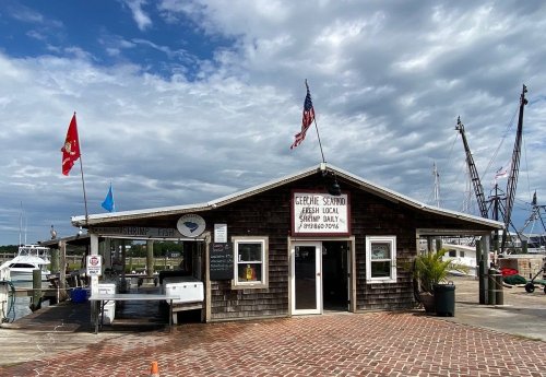 A rustic seafood market with flags, located by a marina under a cloudy sky. Boats are visible in the background.