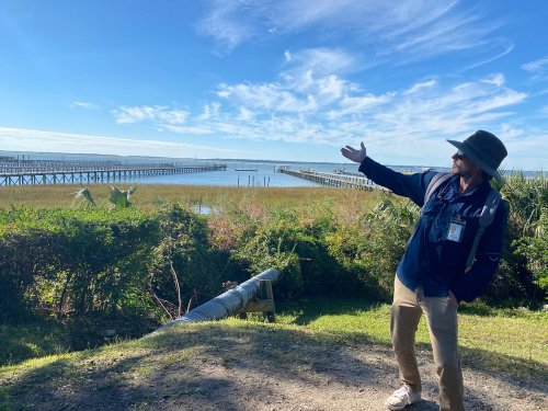 A person in a hat gestures towards a scenic view of a pier and water under a clear blue sky.