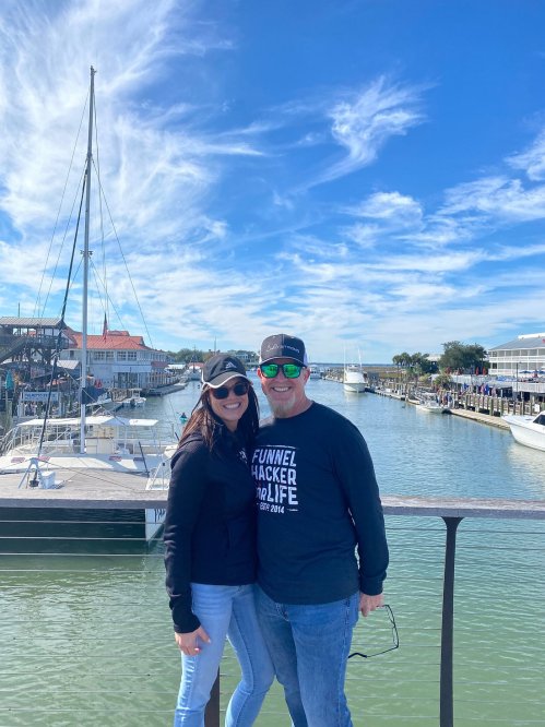 A couple stands on a dock with boats in the background, smiling under a clear blue sky.