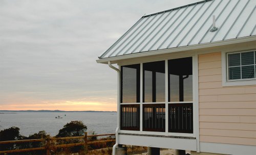 A coastal house with a metal roof and screened porch, overlooking a calm bay at sunset.