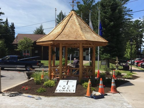 A wooden gazebo with a sign reading "Lest We Forget," surrounded by flowers and construction cones.